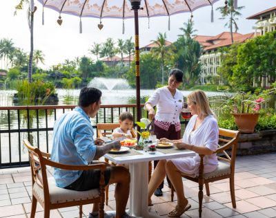 Family dining outdoors near a lake, served by a waitress. Lush greenery and water fountain in the background. Relaxed and cheerful atmosphere.