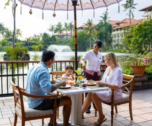 Family dining outdoors near a lake, served by a waitress. Lush greenery and water fountain in the background. Relaxed and cheerful atmosphere.