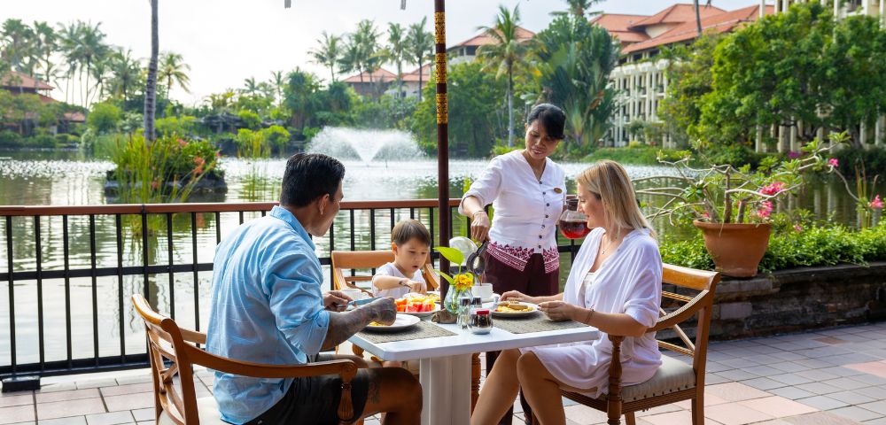 Family dining outdoors near a lake, served by a waitress. Lush greenery and water fountain in the background. Relaxed and cheerful atmosphere.