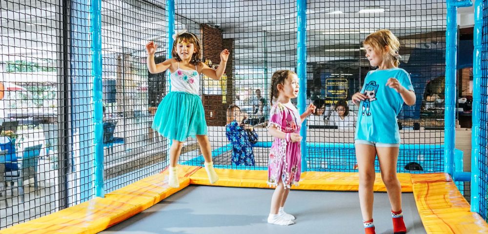 Three young children are joyfully jumping on a trampoline in a vibrant indoor play area. They are surrounded by safety netting, with colorful decor.