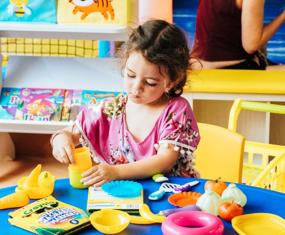 A young girl in a pink dress plays with colorful toy food on a blue table, focused and engaged. Bright books and a yellow chair are in the background.