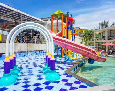 Colorful water playground with a curved red slide and vibrant arches spraying water. Kids play in sunlit, checkered-pool area, creating a lively atmosphere.