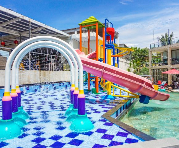 Colorful water playground with a curved red slide and vibrant arches spraying water. Kids play in sunlit, checkered-pool area, creating a lively atmosphere.