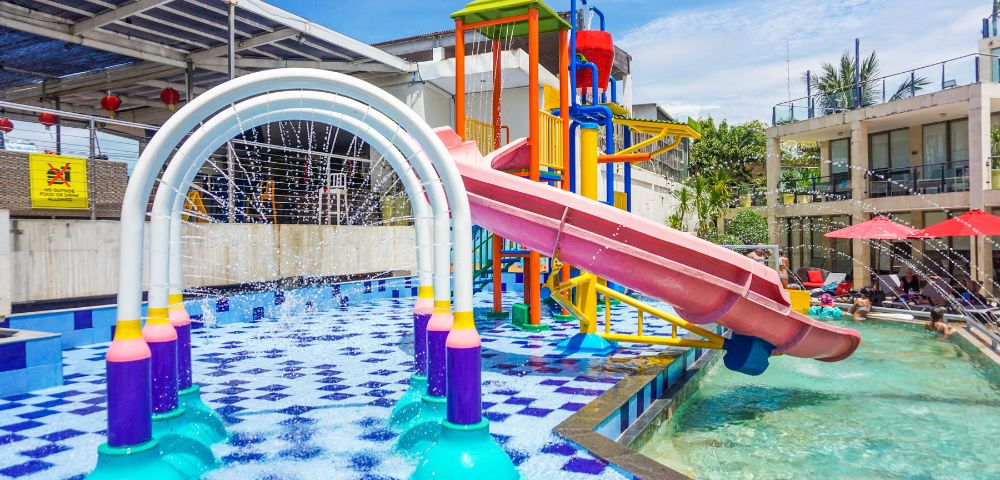 Colorful water playground with a curved red slide and vibrant arches spraying water. Kids play in sunlit, checkered-pool area, creating a lively atmosphere.