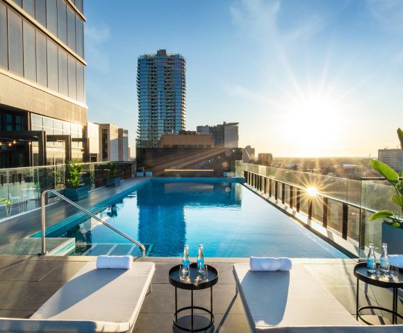 A rooftop infinity pool at sunset, bordered by modern glass railings. Lounge chairs and tables with water bottles in the foreground, cityscape in the background.