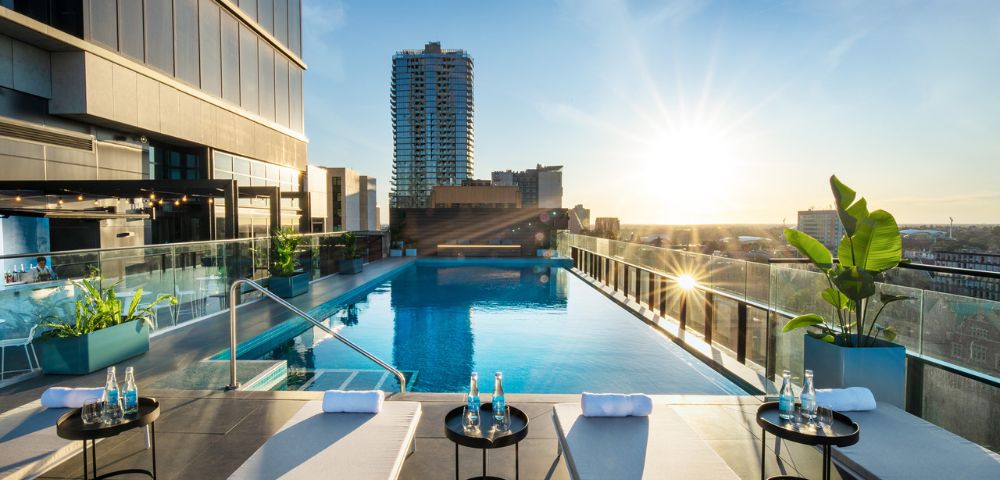 A rooftop infinity pool at sunset, bordered by modern glass railings. Lounge chairs and tables with water bottles in the foreground, cityscape in the background.