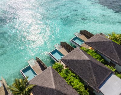 Aerial view of luxury overwater bungalows with thatched roofs, each featuring a private pool, surrounded by clear turquoise ocean and lush green foliage.