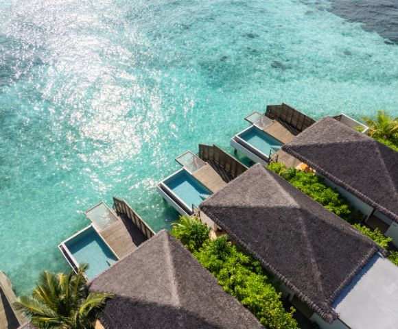 Aerial view of luxury overwater bungalows with thatched roofs, each featuring a private pool, surrounded by clear turquoise ocean and lush green foliage.