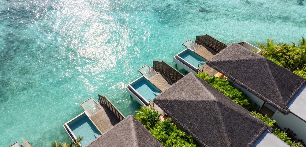 Aerial view of luxury overwater bungalows with thatched roofs, each featuring a private pool, surrounded by clear turquoise ocean and lush green foliage.