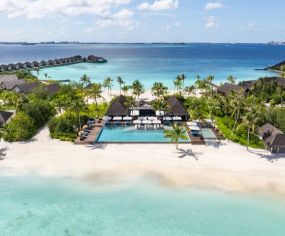 Aerial view of a tropical beach resort with a central pool, surrounded by palm trees and thatched-roof villas. Overwater bungalows extend into the azure ocean.