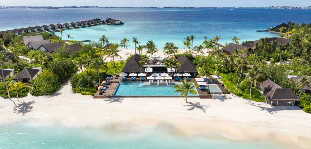 Aerial view of a tropical beach resort with a central pool, surrounded by palm trees and thatched-roof villas. Overwater bungalows extend into the azure ocean.