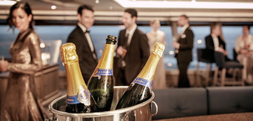 Elegantly dressed people socializing in a luxurious setting with a focus on an ice bucket filled with champagne bottles, conveying a festive mood.