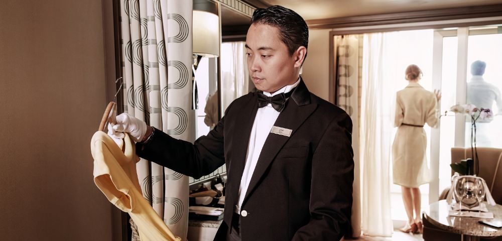 A butler in a tuxedo holds a yellow dress on a hanger in an elegant room. A woman stands by the window, looking out. The scene conveys luxury.
