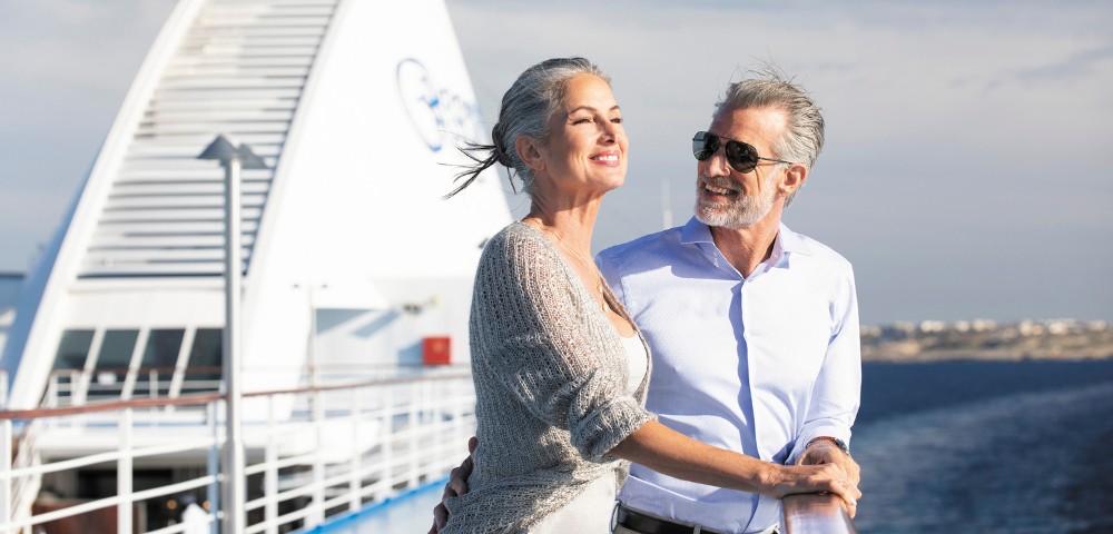 Senior couple on a cruise ship deck, smiling and enjoying the ocean view under a clear blue sky. Their relaxed posture conveys joy and contentment.