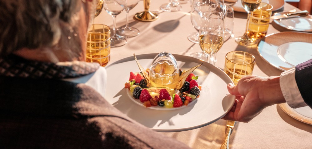 A person is served a sophisticated dessert with fresh berries and an elegant centerpiece on a white plate. The setting is a formal table with numerous empty wine glasses, amber glassware, and shiny cutlery, creating an upscale dining ambiance.