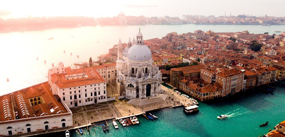 Aerial view of Venice with the prominent dome of Santa Maria della Salute framed by historic buildings and canals. The sun casts a warm glow over the scene.