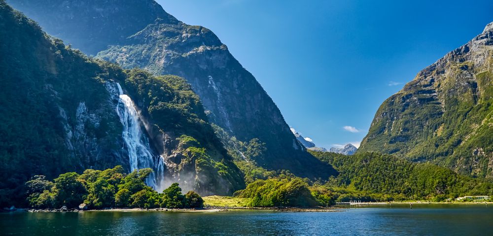 A serene landscape of a waterfall cascading down lush green mountains into a tranquil lake, under a clear blue sky, evoking a sense of peace and natural beauty.