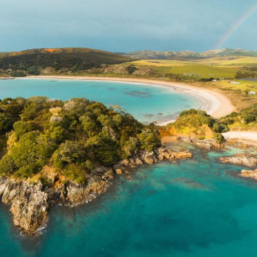 Aerial view of a serene coastline, featuring turquoise waters, a sandy beach, lush green hills, and a faint rainbow under a clear sky.