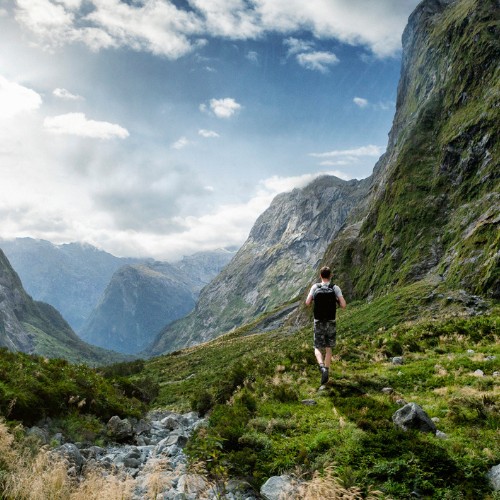 A person hikes through a lush, green mountain valley under a partly cloudy sky. Towering cliffs surround the scene, conveying adventure and solitude.