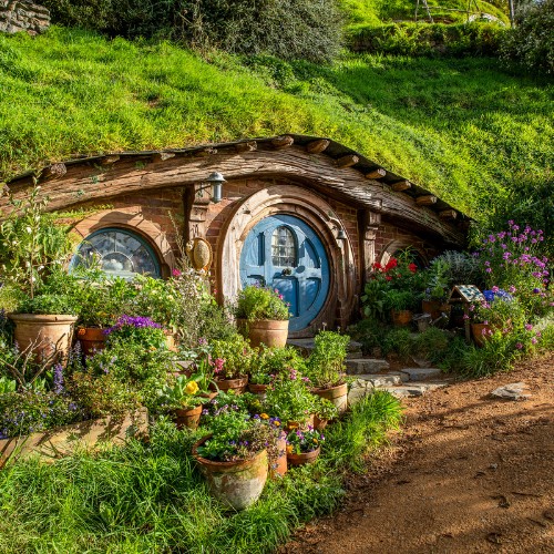 A cozy hobbit-style home with a round blue door is nestled in a grassy hillside, surrounded by vibrant flowers and terracotta pots. The scene feels serene and inviting.