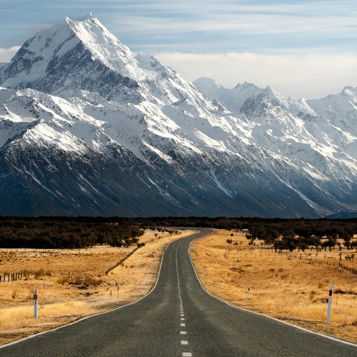A straight, empty road leads towards majestic snow-capped mountains under a clear sky, flanked by golden fields, conveying peace and grandeur.
