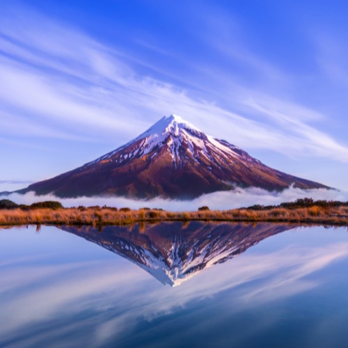 Snow-capped mountain reflecting in a tranquil lake below a clear blue sky with wispy clouds. Serenity and natural beauty dominate the scene.