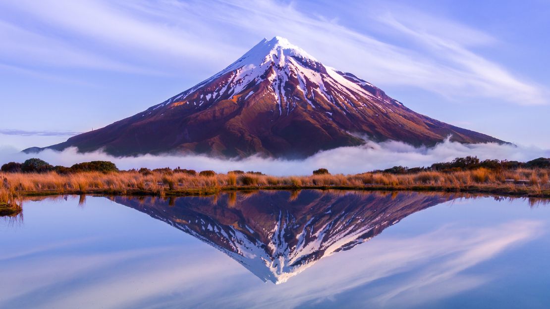 Snow-capped mountain reflecting in a tranquil lake below a clear blue sky with wispy clouds. Serenity and natural beauty dominate the scene.