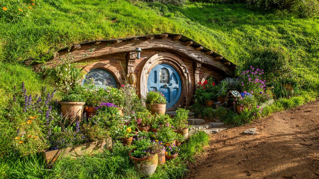 A cozy hobbit-style home with a round blue door is nestled in a grassy hillside, surrounded by vibrant flowers and terracotta pots. The scene feels serene and inviting.