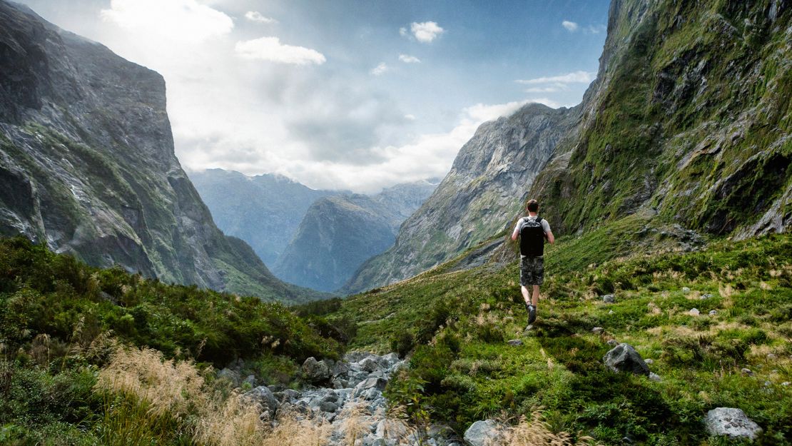 A person hikes through a lush, green mountain valley under a partly cloudy sky. Towering cliffs surround the scene, conveying adventure and solitude.