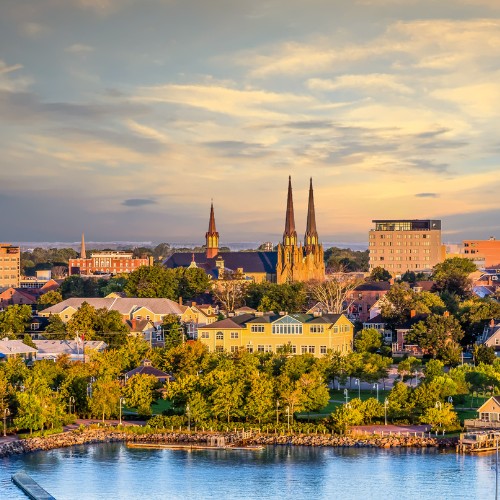 Sunset over Charlottetown, with an emphasis on a cathedral's spires amidst lush trees and colorful buildings, reflecting tranquility near calm waters.