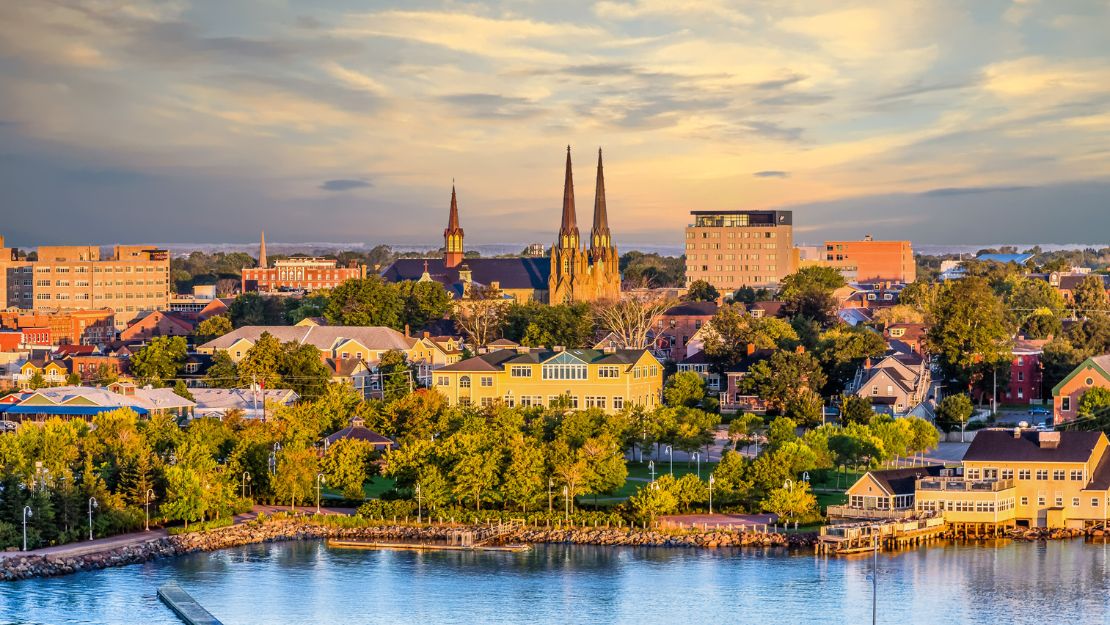 Sunset over Charlottetown, with an emphasis on a cathedral's spires amidst lush trees and colorful buildings, reflecting tranquility near calm waters.