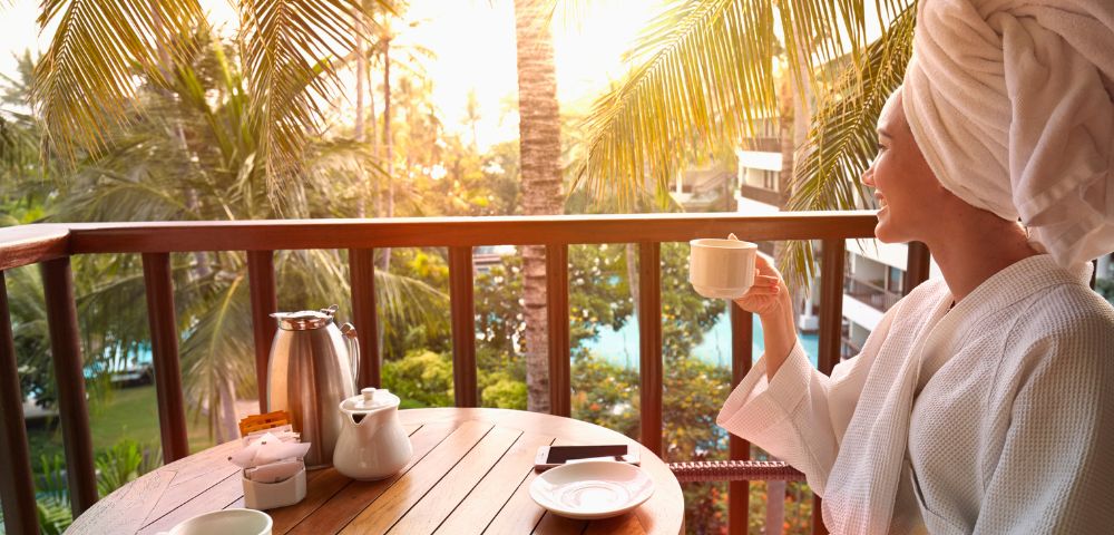 A woman in a bathrobe enjoys morning tea on a balcony surrounded by lush palm trees and a warm golden sunrise.