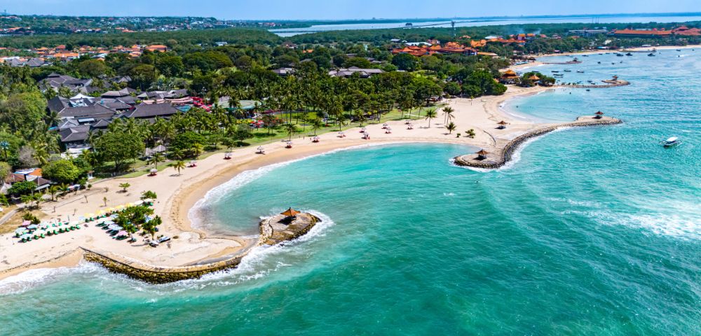 Aerial view of a tropical beach with clear turquoise water, sandy shores, palm trees, and beach huts along the coastline.
