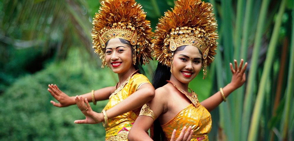 Two dancers dressed in elaborate golden costumes and elaborate headdresses perform gracefully amidst lush green foliage.