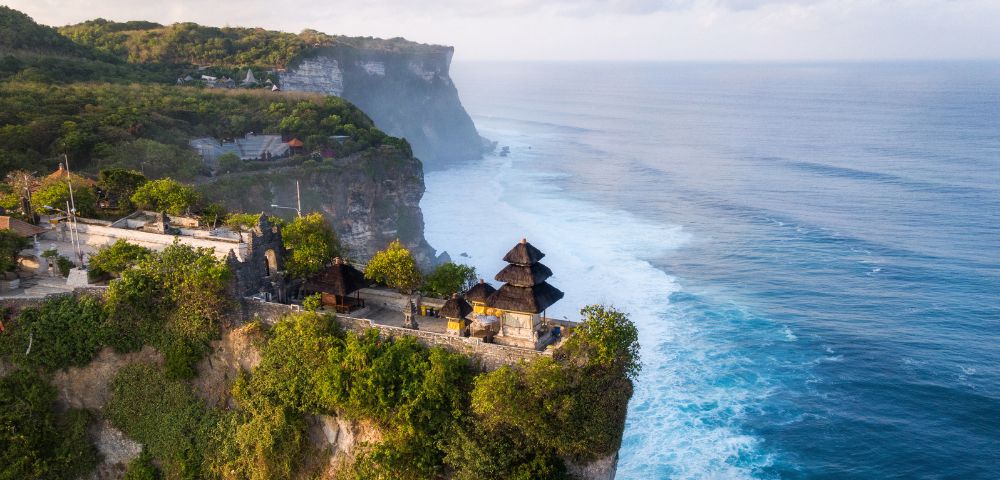 Aerial view of Uluwatu Temple perched on a cliff, surrounded by lush greenery and overlooking the ocean waves in Bali, Indonesia.