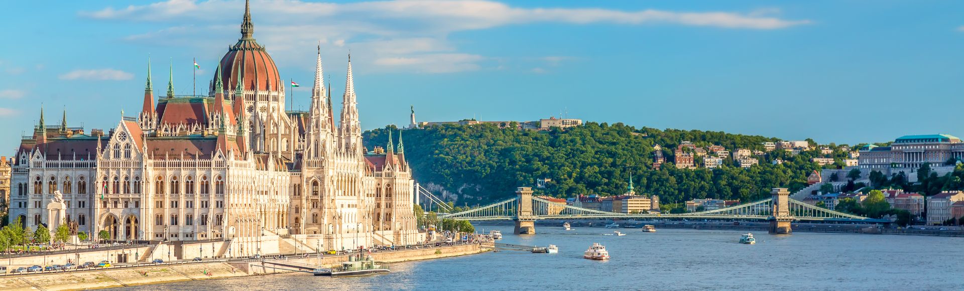 Panoramic view of Budapest's Parliament building along the Danube River, with the Chain Bridge and Buda Hills in the background under a blue sky.