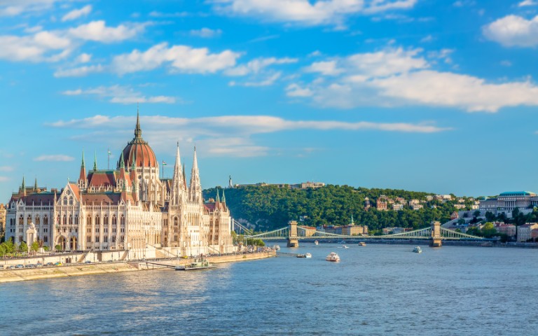 Panoramic view of Budapest's Parliament building along the Danube River, with the Chain Bridge and Buda Hills in the background under a blue sky.