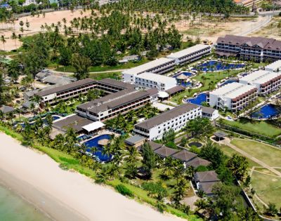 Aerial view of a beachfront resort with multiple modern, white buildings, blue pools, and lush palm trees. The beach is sandy, and the ocean is calm, conveying a relaxing and tropical vibe.