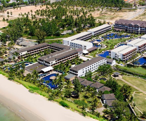 Aerial view of a beachfront resort with multiple modern, white buildings, blue pools, and lush palm trees. The beach is sandy, and the ocean is calm, conveying a relaxing and tropical vibe.