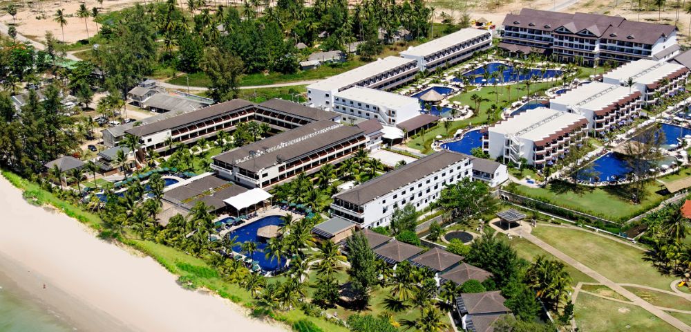 Aerial view of a beachfront resort with multiple modern, white buildings, blue pools, and lush palm trees. The beach is sandy, and the ocean is calm, conveying a relaxing and tropical vibe.