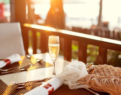 A glass of champagne sparkles on a sunlit outdoor table set for two, with warm, golden light creating a serene and inviting atmosphere.