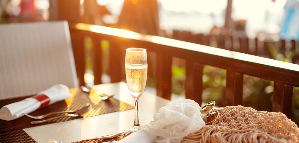 A glass of champagne sparkles on a sunlit outdoor table set for two, with warm, golden light creating a serene and inviting atmosphere.