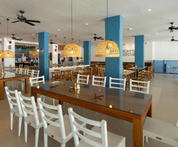 Spacious, modern restaurant interior with wooden tables and white chairs. Woven pendant lights hang from the ceiling, providing a warm, inviting glow.