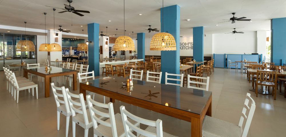 Spacious, modern restaurant interior with wooden tables and white chairs. Woven pendant lights hang from the ceiling, providing a warm, inviting glow.