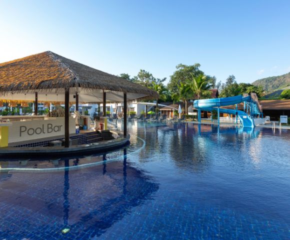 Poolside view with a thatched-roof pool bar and a winding blue water slide, set against lush greenery and a clear blue sky, evoking a relaxing resort vibe.