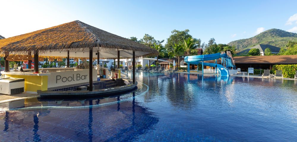 Poolside view with a thatched-roof pool bar and a winding blue water slide, set against lush greenery and a clear blue sky, evoking a relaxing resort vibe.