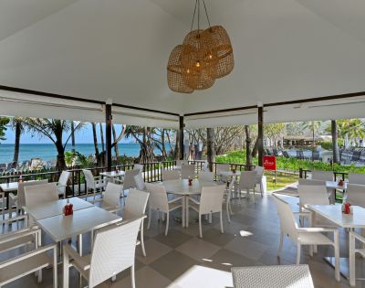 Outdoor restaurant with white wicker furniture and a woven chandelier, overlooking a beach with palm trees and ocean. Bright, tropical, and inviting ambiance.