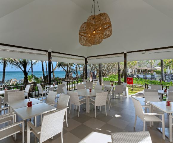 Outdoor restaurant with white wicker furniture and a woven chandelier, overlooking a beach with palm trees and ocean. Bright, tropical, and inviting ambiance.