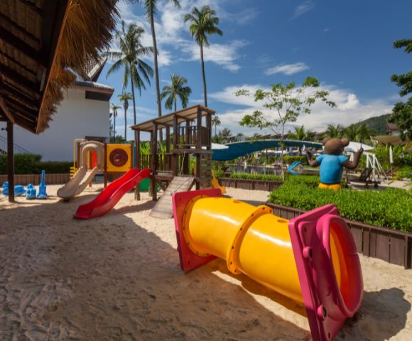 Playground with vibrant slides, climbing structures, and a tunnel on sand. Palm trees and a sunny sky create a lively, tropical atmosphere.