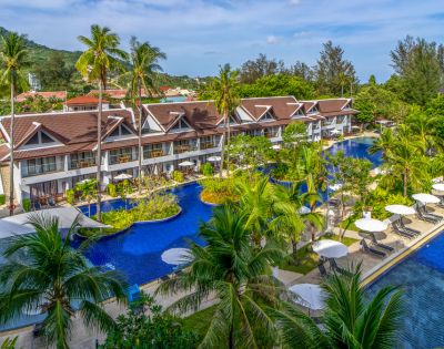 Tropical resort with a series of bungalows beside a winding blue pool, surrounded by palm trees and lush greenery. Lounge chairs and umbrellas line the water.